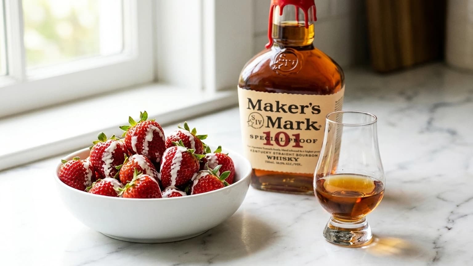 Bowl of fresh strawberries with a cream drizzle beside a Glencairn of bourbon and a bottle of Maker's Mark on a marble counter in window light