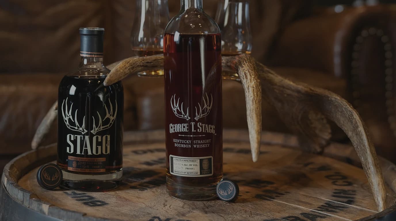 George T. Stagg and Stagg Jr. bottles on a whiskey barrel with a mounted antler and Glencairn glasses behind, leather armchair in soft focus — the Buffalo Trace Antique Collection Stagg that took Best in Show at the 2026 ASCOT Awards