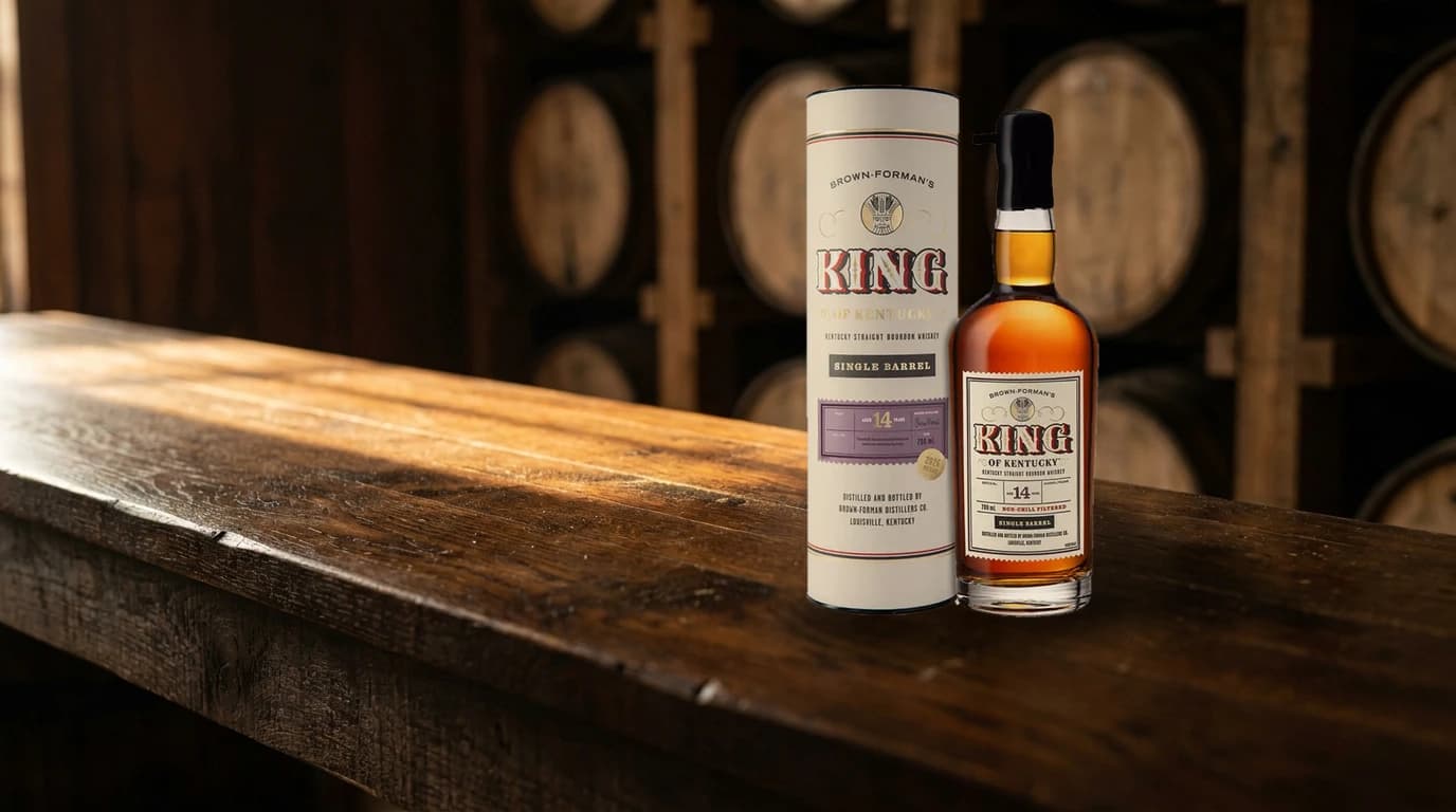 A bottle of Brown-Forman's King of Kentucky 14 Year Single Barrel 2026 bourbon with its cream canister and black wax-dipped neck, standing on a dark oak bar top in warm side light with rickhouse barrels behind