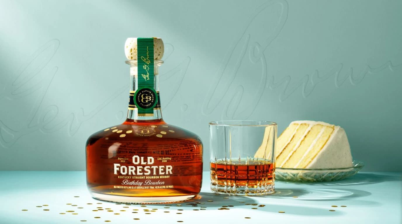 Old Forester 2026 Birthday Bourbon bottle with its green George Garvin Brown neck ribbon beside a rocks glass pour and a slice of white layer cake on a pale blue backdrop